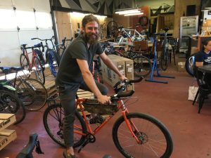 Bike mechanic Caleb Murray smiles for the camera while sitting on his bicycle. He says he hopes to see a junior mechanic come in that he can train as his workload gets bigger. Photo: Brock Ormond, QNet News.